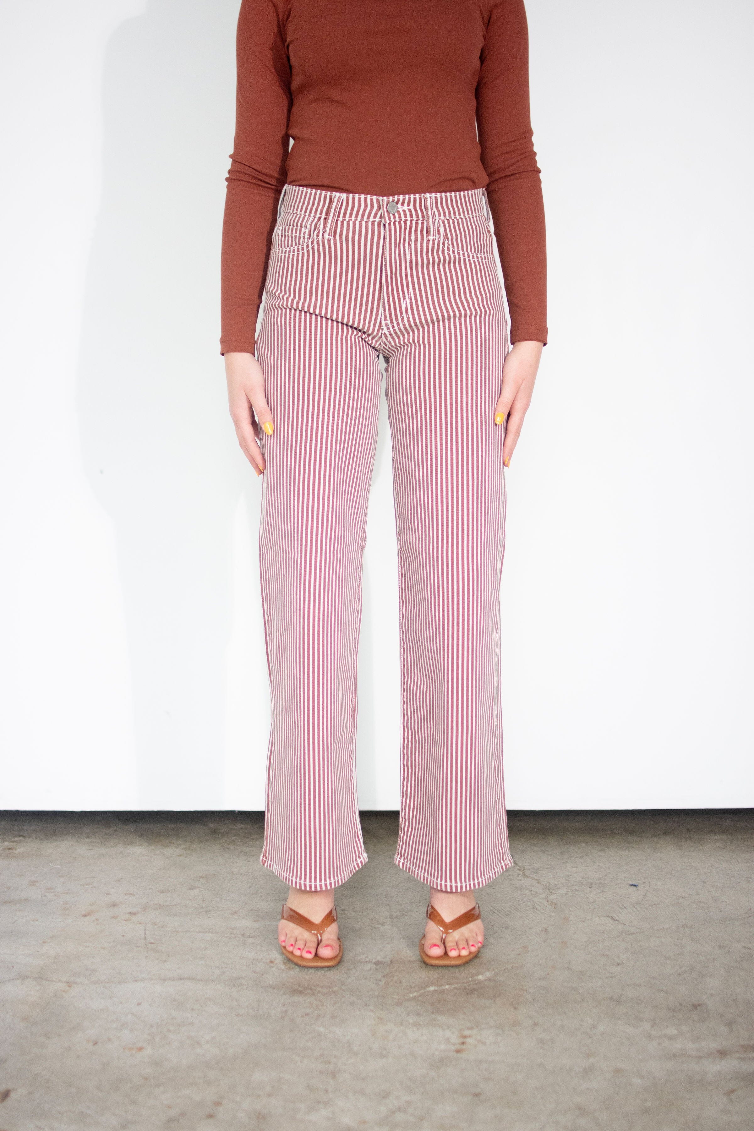 A model wearing pink and white vertically striped wide-leg jeans, paired with a rust-brown long-sleeve top and brown sandals, standing against a plain white wall.