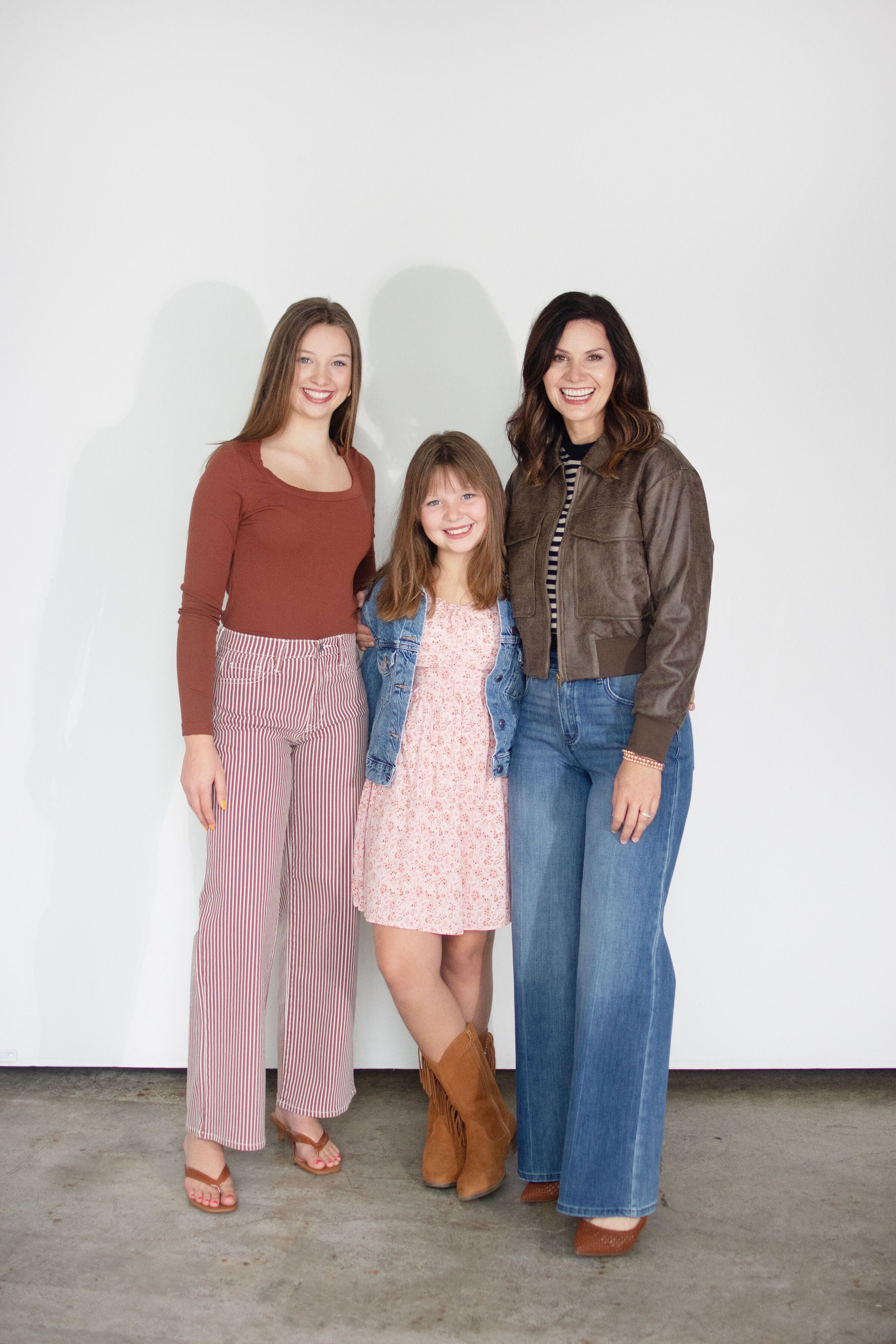 Three women posing together against a plain white wall, showcasing different styles of wide-leg jeans. The woman on the left wears pink ribbed wide-leg jeans with a rust-brown long-sleeve top.