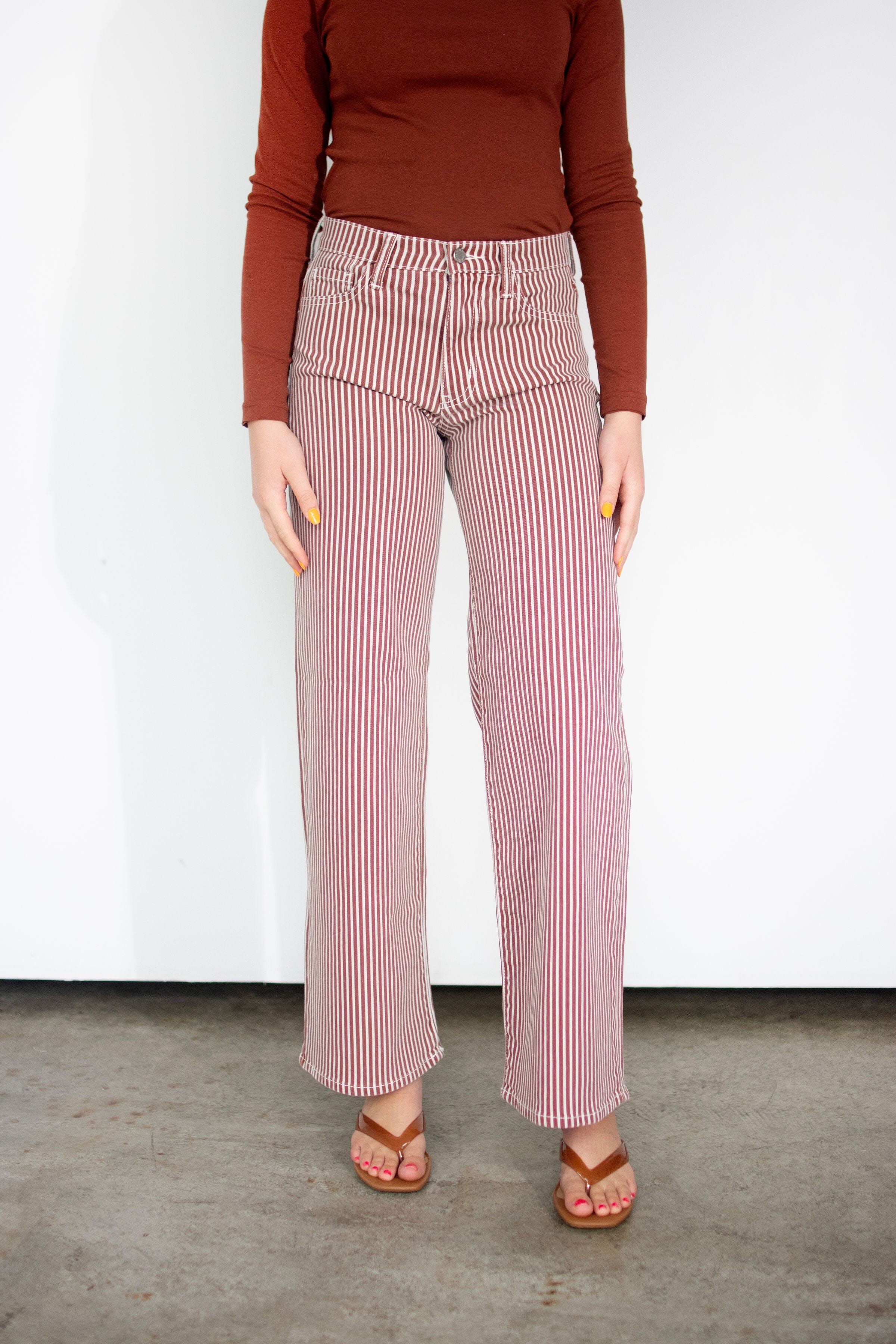 A person wearing high-waisted, wide-leg striped pants in pink and white, paired with a rust-brown long-sleeve top and brown sandals, standing against a white wall.