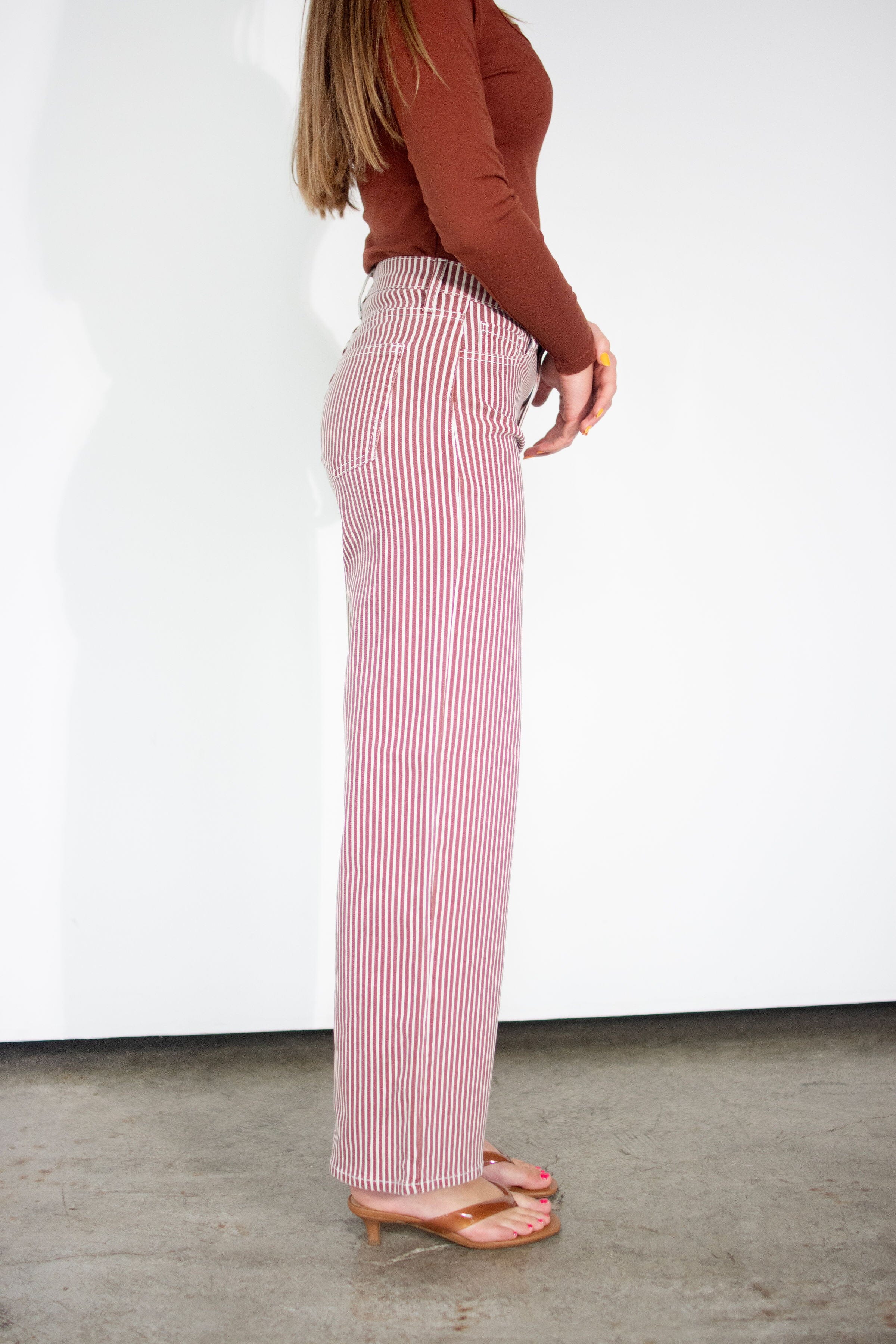 A side-profile view of a model wearing pink and white vertically striped wide-leg pants, paired with a rust-brown long-sleeve top and tan heeled sandals, standing against a white wall.