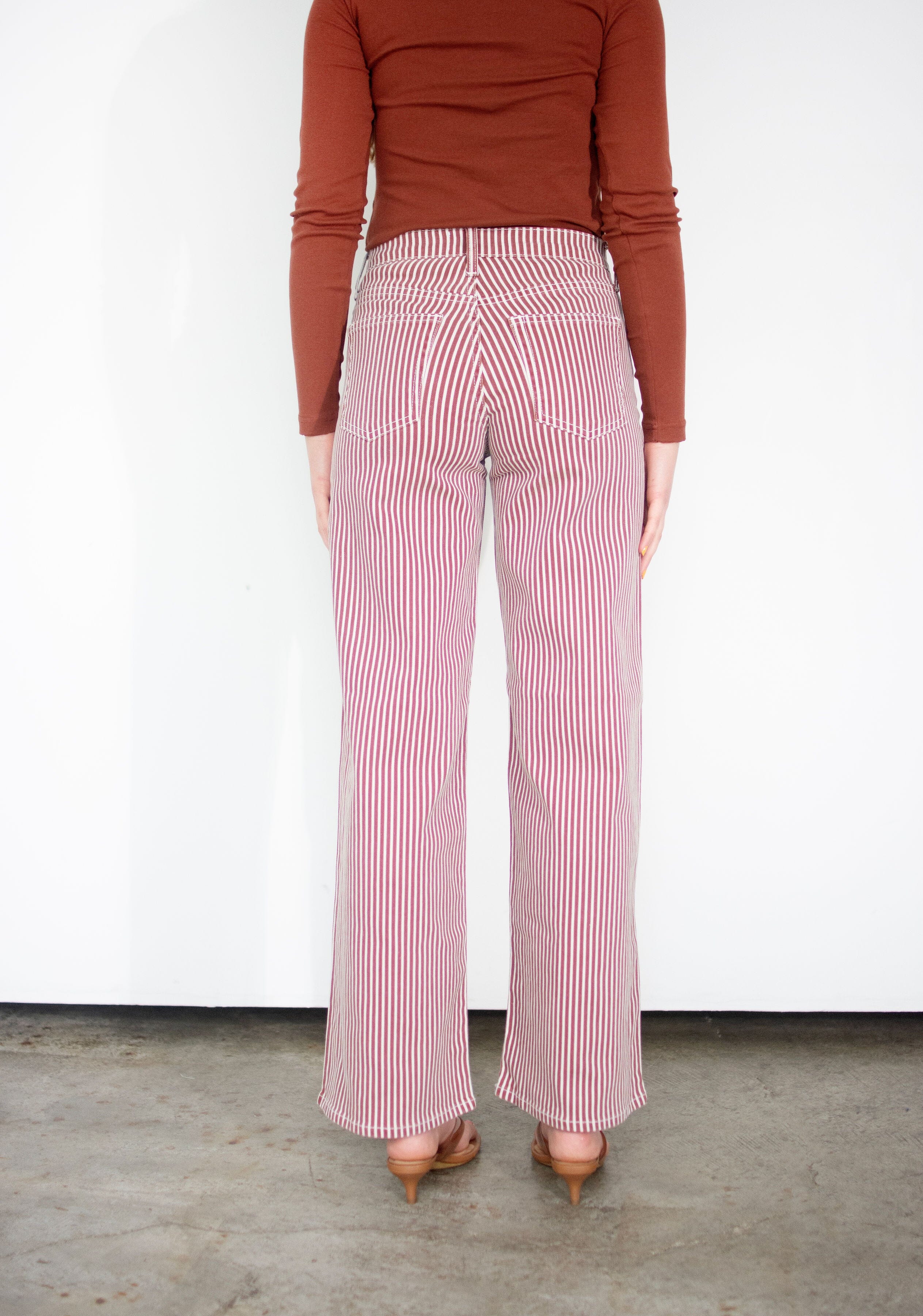 A person shown from the back wearing spice brown and white vertically striped wide-leg pants, a rust-brown long-sleeve top, and tan pointed-toe heels against a plain white wall and concrete floor.